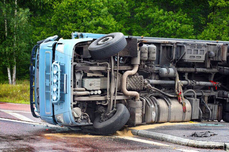A light blue tractor trailer truck has rolled onto its side at the edge of a road.